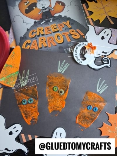 Children’s carrot art made with orange paint and googly eyes is displayed on black paper next to ghost cutouts and the book Creepy Carrots! by Aaron Reynolds. This fun display, perfect for Halloween Crafts for Kids, includes the @GLUEDTOMYCRAFTS watermark.