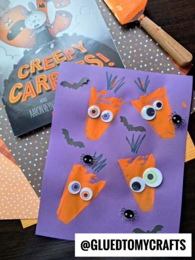 Purple paper with orange carrot shapes, googly eyes, and drawn green stems, plus small paper bats and spiders; a Creepy Carrots! book, a pie server stamped with fun details, spatula, and polka dot paper are also visible. @GLUEDTOMYCRAFTS is at the bottom.