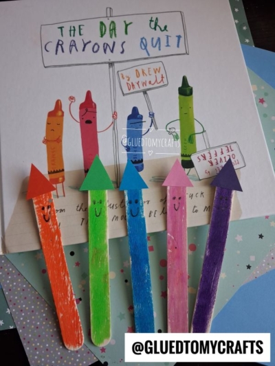 Colorful craft sticks painted as crayons lie on a book titled The Day The Crayons Quit, with each stick decorated with a smiling face and a matching paper tip—making this scene perfect for those looking for creative summer craft ideas. An Instagram handle @GLUEDTOMYCRAFTS appears twice.