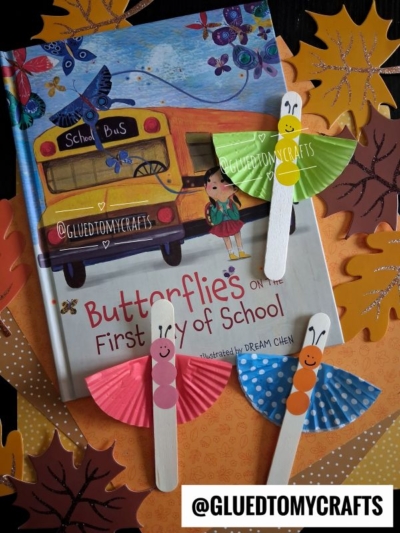 A children’s book titled Butterflies on the First Day of School is surrounded by paper leaves and three colorful cupcake liner butterfly crafts made from cupcake liners and popsicle sticks. @GLUEDTOMYCRAFTS is labeled at the bottom.