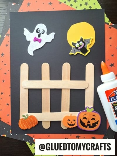 Get creative this spooky season with one of the best Halloween Crafts for Kids—a festive scene featuring a popsicle stick fence, foam pumpkin and ghost stickers, a bat soaring by a yellow moon, and a trusty bottle of Elmers glue, all set against starry orange and black paper. @GLUEDTOMYCRAFTS is at the bottom.