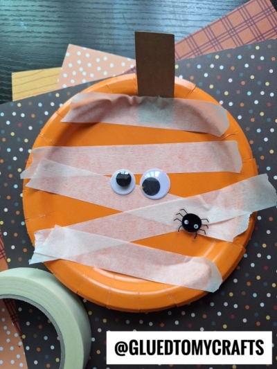 An orange paper plate with googly eyes, a small plastic spider, and tan painter’s tape strips, resembling a mummy, on top of polka dot paper and crafting materials—perfect for anyone seeking creative painter’s tape craft ideas.