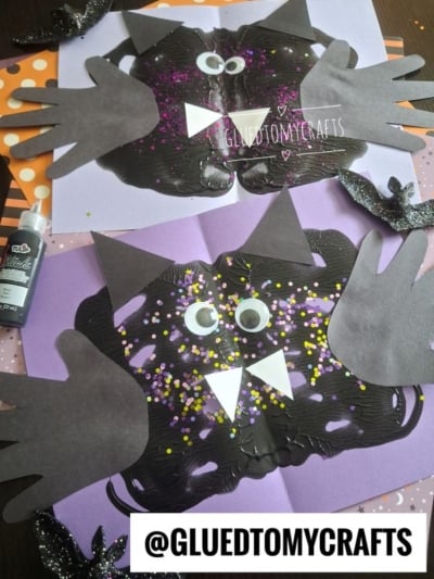 Two paper bat crafts with handprint wings, googly eyes, and glitter on purple paper. Black paint forms the bat bodies, with paper triangles for fangs and ears. Scrapbook paper and craft supplies are in the background.