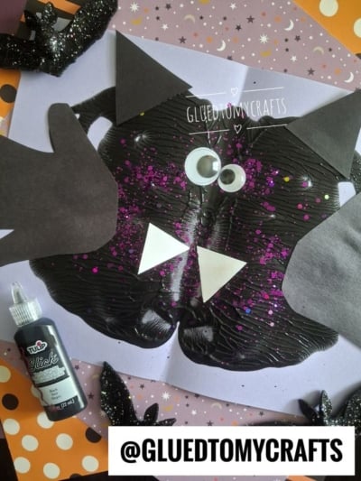 A kids bat craft made with black paint, googly eyes, glitter, and paper shapes on purple paper. A black glittery bat cutout and black fabric paint are nearby. Text: @GLUEDTOMYCRAFTS.