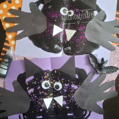 Two black paper bats with handprint wings, googly eyes, and white fangs on purple and orange polka-dot backgrounds, surrounded by glittery cut-out bats and crafting supplies. @GLUEDTOMYCRAFTS text at the bottom.