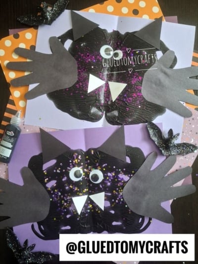 Two black paper bats with handprint wings, googly eyes, and white fangs on purple and orange polka-dot backgrounds, surrounded by glittery cut-out bats and crafting supplies. @GLUEDTOMYCRAFTS text at the bottom.