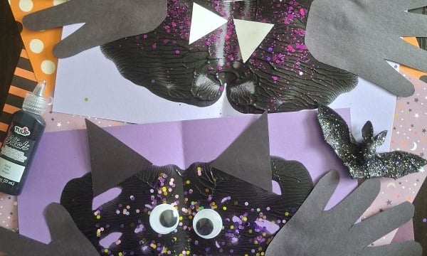 Two black paper bats with handprint wings, googly eyes, and white fangs on purple and orange polka-dot backgrounds, surrounded by glittery cut-out bats and crafting supplies. @GLUEDTOMYCRAFTS text at the bottom.