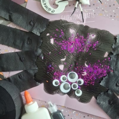 Paper handprint spider craft with black paint, googly eyes, purple glitter, glue, and crafting supplies on a decorated background. @GLUEDTOMYCRAFTS logo appears at the bottom.