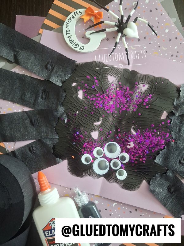 Paper handprint spider craft with black paint, googly eyes, purple glitter, glue, and crafting supplies on a decorated background. @GLUEDTOMYCRAFTS logo appears at the bottom.