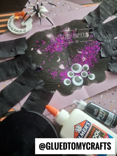 A Halloween spider craft made of black paint, googly eyes, purple glitter, and paper strips. Supplies like glue and a marker are nearby. The text @GLUEDTOMYCRAFTS is on the image.