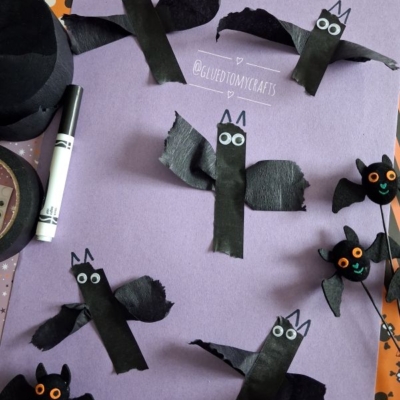 Paper bats made from black paper, googly eyes, and torn tissue wings are arranged on a purple sheet. Halloween-themed craft materials and a marker surround the artwork.
