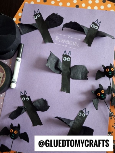 Paper bats made from black paper, googly eyes, and torn tissue wings are arranged on a purple sheet. Halloween-themed craft materials and a marker surround the artwork.