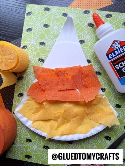 A paper gnome craft in progress, made with orange and yellow crepe paper on a white base, glue bottle nearby. Perfect for Halloween Crafts for Kids, the project rests on green paper with a spider web pattern underneath.