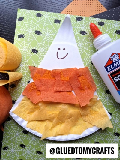 A candy corn-themed craft made with tissue paper, a paper plate, and glue sits on a spiderweb-patterned background. A smiley face is drawn on the white tip. Crepe paper and glue are nearby.