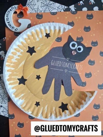 A Halloween craft featuring a paper plate moon crescent, a black cat made from a traced hand, black stars, and an orange background with black cat faces. Text @GLUEDTOMYCRAFTS is shown.