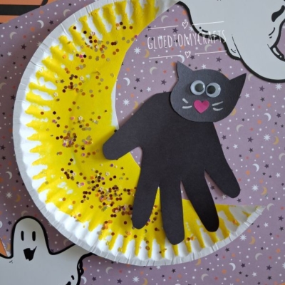 A Halloween craft featuring a black cat made from a handprint, sitting on a yellow crescent moon decorated with glitter, surrounded by paper ghost cutouts and Halloween-themed paper. @GLUEDTOMYCRAFTS is written at the bottom.