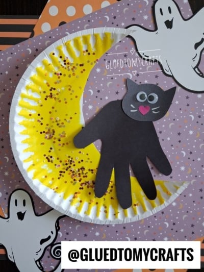 A Halloween craft featuring a black cat made from a handprint, sitting on a yellow crescent moon decorated with glitter, surrounded by paper ghost cutouts and Halloween-themed paper. @GLUEDTOMYCRAFTS is written at the bottom.