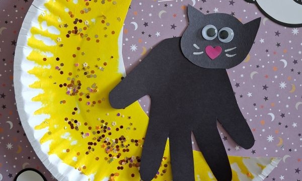 A Halloween craft featuring a black cat made from a handprint, sitting on a yellow crescent moon decorated with glitter, surrounded by paper ghost cutouts and Halloween-themed paper. @GLUEDTOMYCRAFTS is written at the bottom.