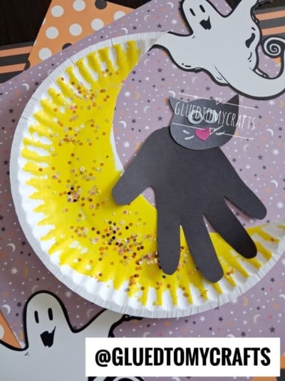 A Halloween craft with a yellow, glittery crescent moon on a paper plate, a black paper cat silhouette, and ghost decorations on a purple background. Text @GLUEDTOMYCRAFTS appears at the bottom.