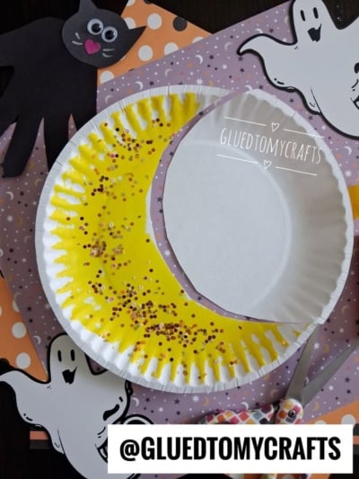 A paper plate craft features a yellow crescent moon with brown glitter, surrounded by paper ghost and bat decorations on a purple polka-dot background. Scissors and the handle of a glue stick are also visible.