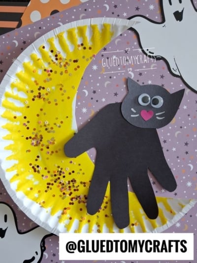 A paper plate craft features a yellow crescent moon with gold glitter and a black cat made from a handprint and paper shapes, set on a Halloween-themed background.