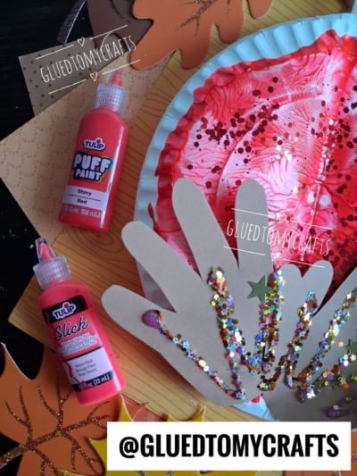 Paper plate craft with red paint, glitter, and handprint cutouts, plus bottles of red puff and slick paint. Autumn leaf cutouts and the handle @GLUEDTOMYCRAFTS are visible.