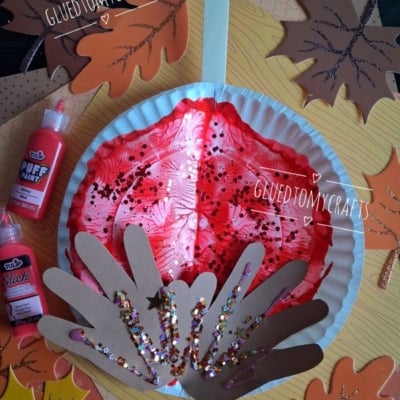 A fall craft with a red painted paper plate, brown handprint cutouts, and glitter, surrounded by paper leaves, paint bottles, and the text @GLUEDTOMYCRAFTS..