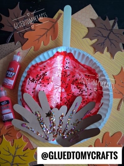 A fall craft with a red painted paper plate, brown handprint cutouts, and glitter, surrounded by paper leaves, paint bottles, and the text @GLUEDTOMYCRAFTS..