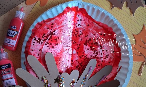 A fall craft with a red painted paper plate, brown handprint cutouts, and glitter, surrounded by paper leaves, paint bottles, and the text @GLUEDTOMYCRAFTS..
