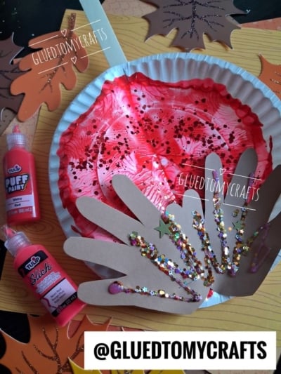 A red, glittery paper plate craft with brown paper handprints, glitter, and paint sits among colorful paper leaves. Paint bottles and the username @GLUEDTOMYCRAFTS are also visible.