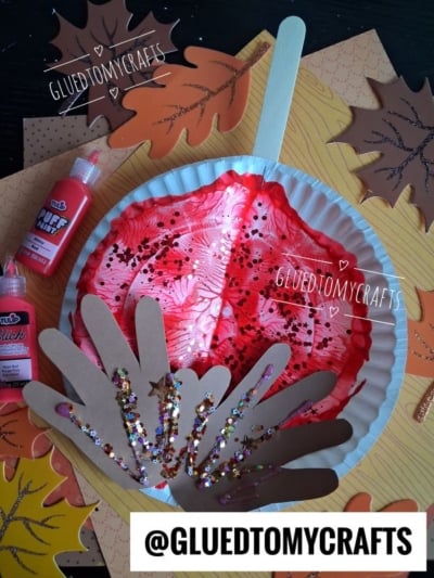 A paper plate craft shaped like a fall tree, decorated with red and pink paint, brown paper handprints for branches, and glitter. Bottles of craft glue and sparkly leaf decorations surround the plate.