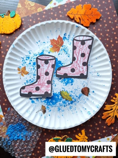 Paper plate rain puddle craft featuring blue paint, fall leaves, and cutouts of pink polka-dot rain boots on a brown background with autumn decorations. @GLUEDTOMYCRAFTS text label at the bottom.