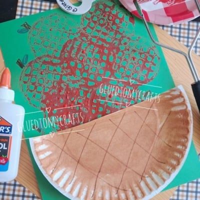 A paper plate painted brown and folded is on a table with a green paper featuring a red apple print, glue, and a plaid napkin. A paint roller and red paint-covered plate are also visible.