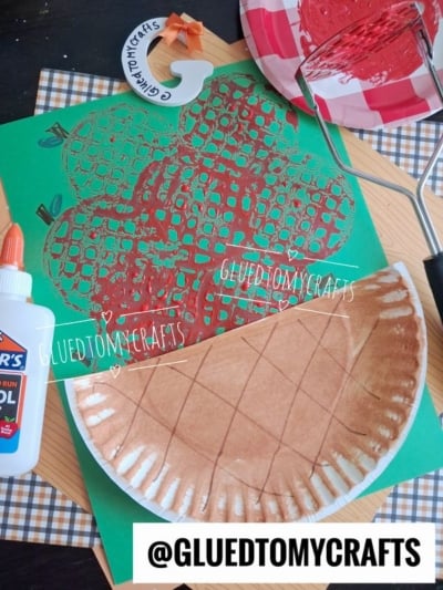 A paper plate painted brown and folded is on a table with a green paper featuring a red apple print, glue, and a plaid napkin. A paint roller and red paint-covered plate are also visible.