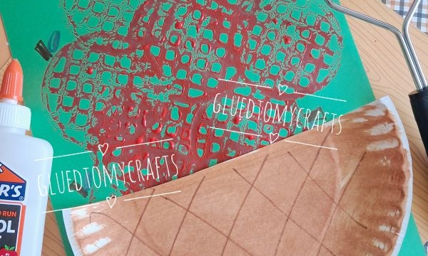 A paper plate painted brown and folded is on a table with a green paper featuring a red apple print, glue, and a plaid napkin. A paint roller and red paint-covered plate are also visible.