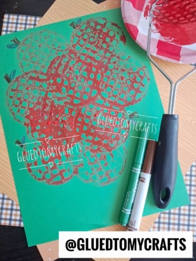A green paper shows a red apple print pattern made with a textured roller, next to a brown and green marker, a paint roller, and a red paint-covered plate. Text reads @GLUEDTOMYCRAFTS..