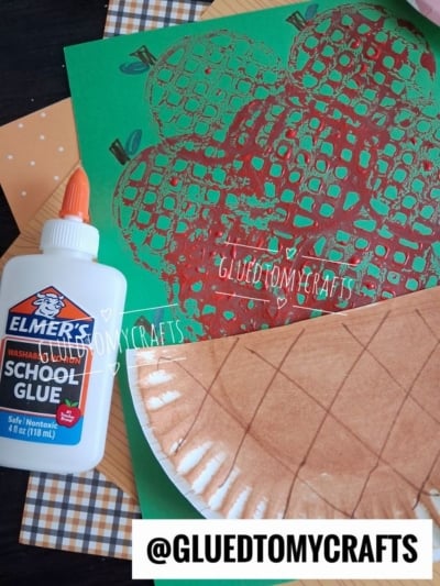 A green paper with a red apple print, a bottle of Elmers school glue, a brown paper decorated like a pie crust, and patterned papers are arranged on a table. @GLUEDTOMYCRAFTS watermark is visible.