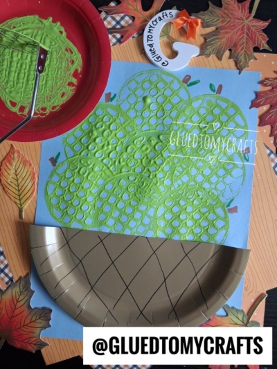 A kids craft project shows a painted green acorn top made with a bubble wrap paint print and a brown paper plate as the acorn bottom, surrounded by fake leaves, craft supplies, and a fun potato masher stamped apple for an extra pop of color.