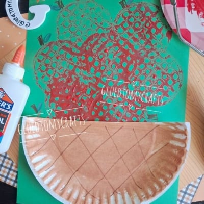 A green paper with a red apple print, a brown paper plate craft resembling a pie, a bottle of Elmers glue, and a paint palette are arranged on a checkered surface. @GLUEDTOMYCRAFTS watermark is visible.