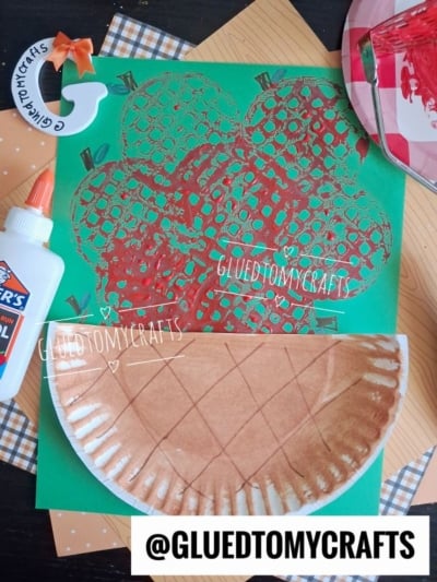 A green paper with a red apple print, a brown paper plate craft resembling a pie, a bottle of Elmers glue, and a paint palette are arranged on a checkered surface. @GLUEDTOMYCRAFTS watermark is visible.