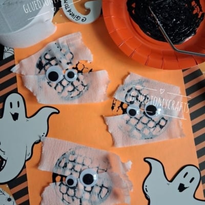 Three ghost crafts with googly eyes and white tissue strips on orange paper, surrounded by ghost cutouts, a black-painted plate, and a paintbrush. The handle @GLUEDTOMYCRAFTS is repeated on the image.