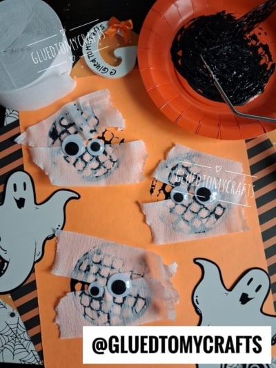 Three ghost crafts with googly eyes and white tissue strips on orange paper, surrounded by ghost cutouts, a black-painted plate, and a paintbrush. The handle @GLUEDTOMYCRAFTS is repeated on the image.