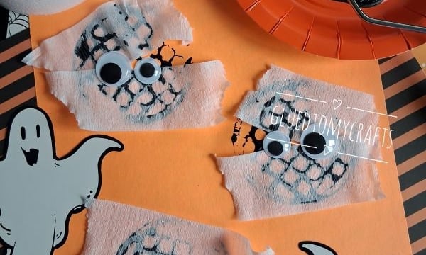 Three ghost crafts with googly eyes and white tissue strips on orange paper, surrounded by ghost cutouts, a black-painted plate, and a paintbrush. The handle @GLUEDTOMYCRAFTS is repeated on the image.