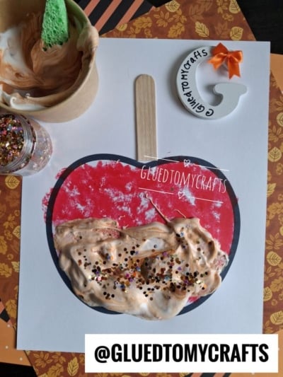 A delightful paper apple craft features brown cream and glitter to resemble a puffy paint caramel apple, complete with a wooden stick for a stem, a paintbrush, and a paint cup set against an autumn-themed background. Text reads @GLUETOMYCRAFTS and GLUETOMYCRAFTS on the apple.