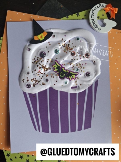 Handmade Halloween cupcake card featuring a whimsical witch cupcake with puffy white frosting, googly eyes, a bat sticker, and a tiny witch hat dusted in glitter. This playful puffy paint witch cupcake design pops against purple and orange paper, finished with a charming craft crescent moon in the corner.