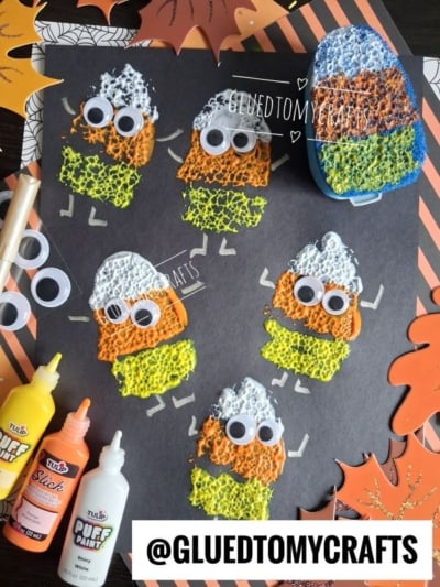 Colorful sponge-painted candy corn characters with googly eyes and pipe cleaner arms are displayed on black paper, making this a fun idea for Halloween Crafts for Kids. Puffy paint bottles and autumn leaf decorations surround the artwork. Instagram handle @GLUEDTOMYCRAFTS is shown.