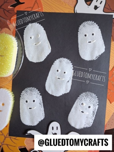 Black paper with six white ghost shapes made from paint and decorated with simple eyes and mouths, perfect for Halloween Crafts for Kids. A ghost-shaped sponge and orange background with white ghosts can be seen around the paper. @GLUETOMYCRAFTS is labeled.