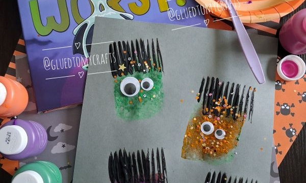 Three monster crafts made with painted sponges, googly eyes, black fork prints, and glitter are on paper, surrounded by art supplies, a Monsters Are The Worst! book, and a plate with a painted sponge.