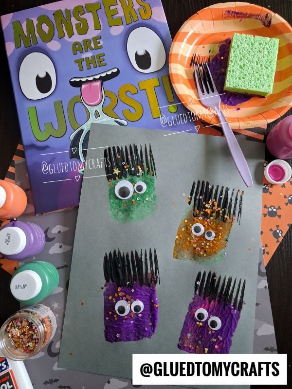 Three monster crafts made with painted sponges, googly eyes, black fork prints, and glitter are on paper, surrounded by art supplies, a Monsters Are The Worst! book, and a plate with a painted sponge.