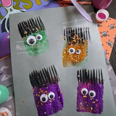 Four colorful monster faces painted on paper with googly eyes and glitter, created using paint and a black plastic fork. Craft supplies and Halloween-themed items are scattered around the background. @GLUEDTOMYCRAFTS is labeled.